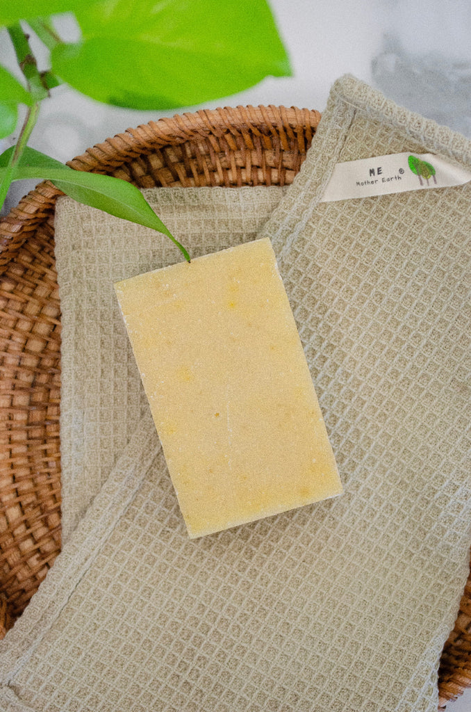 Bar of soap on a textured towel with a wicker basket and green leaves in the background