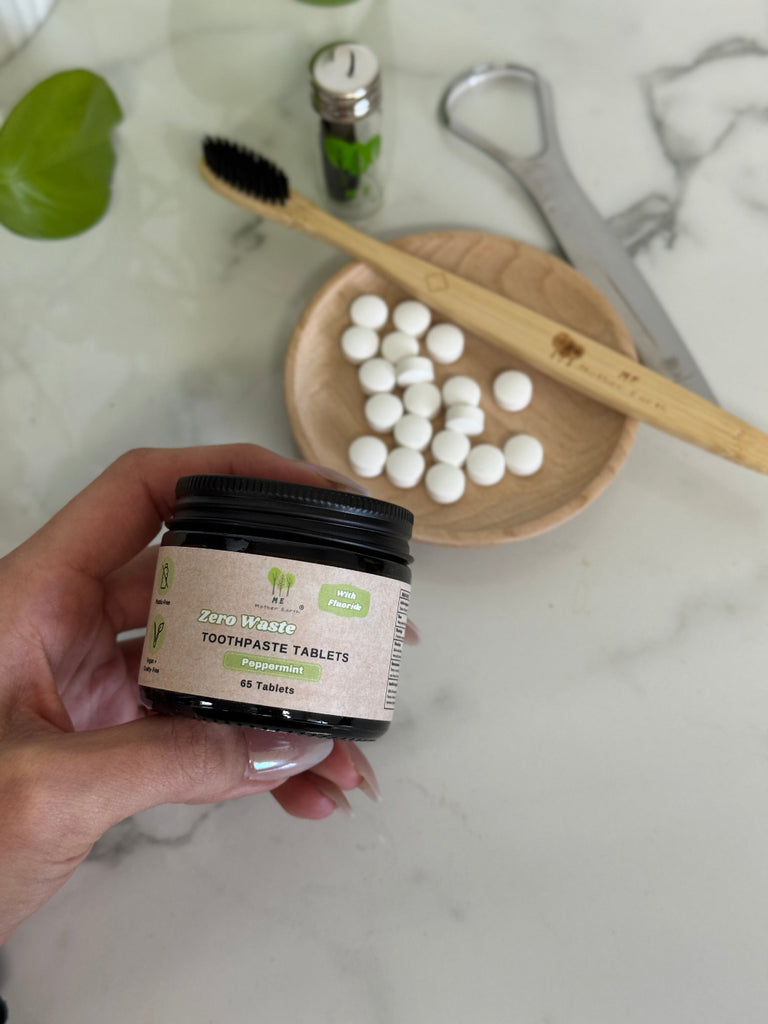 Hand holding a jar of toothpaste tablets with a wooden spoon and tablets on a marble surface.
