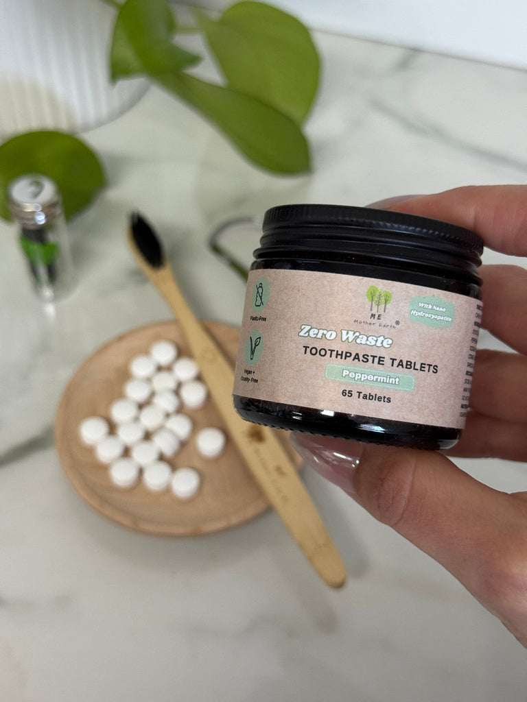 Hand holding a container of zero waste toothpaste tablets with a wooden toothbrush and tablets on a surface.