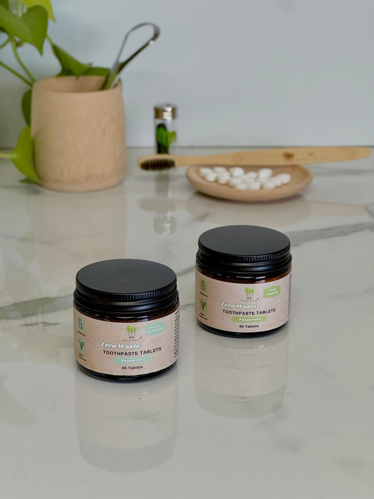 Two jars of toothpaste tablets on a light surface with a plant and wooden items in the background.