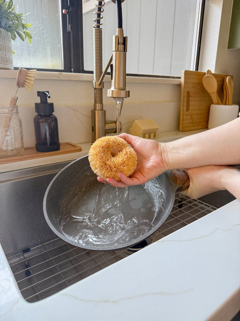 Person holding a natural sponge over a kitchen sink with a window and utensils in the background.