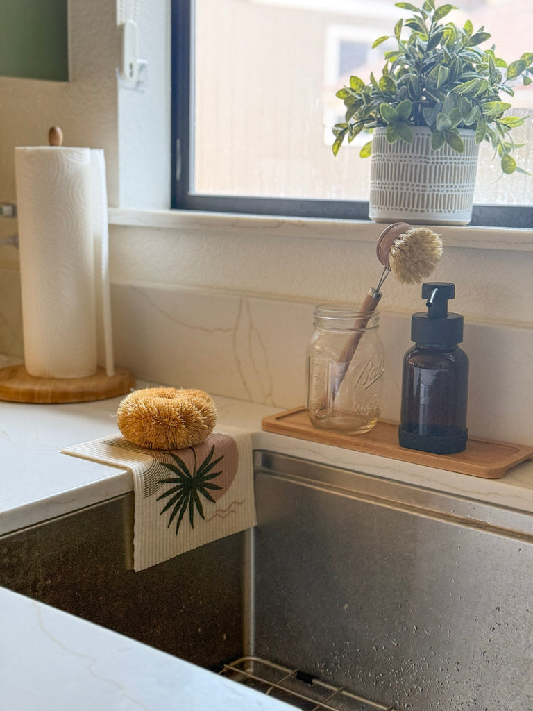 Kitchen sink area with a plant, sponge, and cleaning supplies on a windowsill.